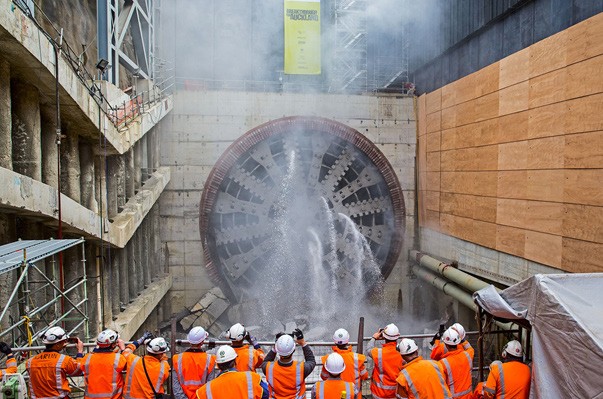 [image] people in hard hats and orange jackets watching tunnel boring machine breakthrough a tunnel